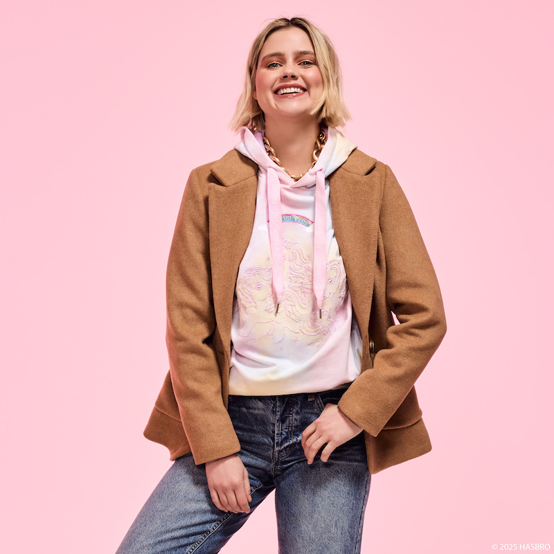 Woman smiling at camera wearing the Loungefly My Little Pony hoodie under a brown jacket with jeans. She stands against a pink background.
