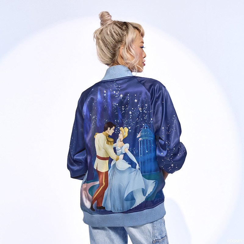 Woman facing away from camera wearing the blue satin Cinderella bomber jacket, featuring an image of Cinderella and Prince Charming on the back of the jacket. 