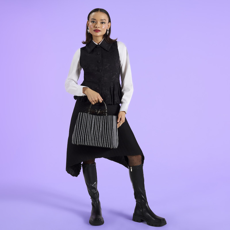 Woman wearing a black sweater over a white long-sleeve top and a black skirt and knee-high boots holding the Loungefly Jack Skellington handbag. She stands against a purple background. 