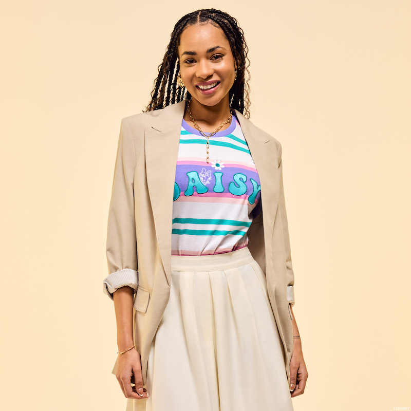 Woman smiling at camera wearing the Loungefly Daisy Duck Tee tucked into a light beige skirt and wearing a beige blazer over it. She stands against a beige background. 