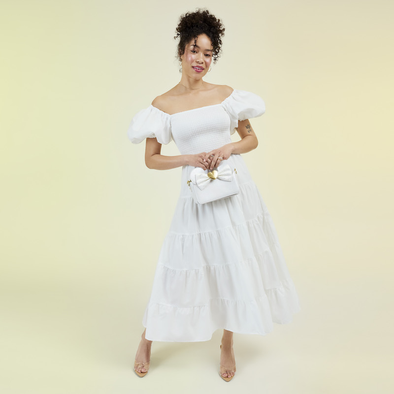Woman wearing a white off-the-shoulder dress and nude heels, holding the Loungefly Minnie Mouse wedding crossbody bag. She stands against a light yellow background. 