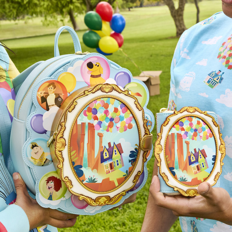 Two people standing outside in a park with a bunch of balloons behind them in the background. They're holding the Loungefly Pixar UP Exclusive 15th Anniversary Carl and Ellie Cameo Mini Backpack and wallet, which features Carl and Ellie's house in Paradise Falls suspended by balloons.