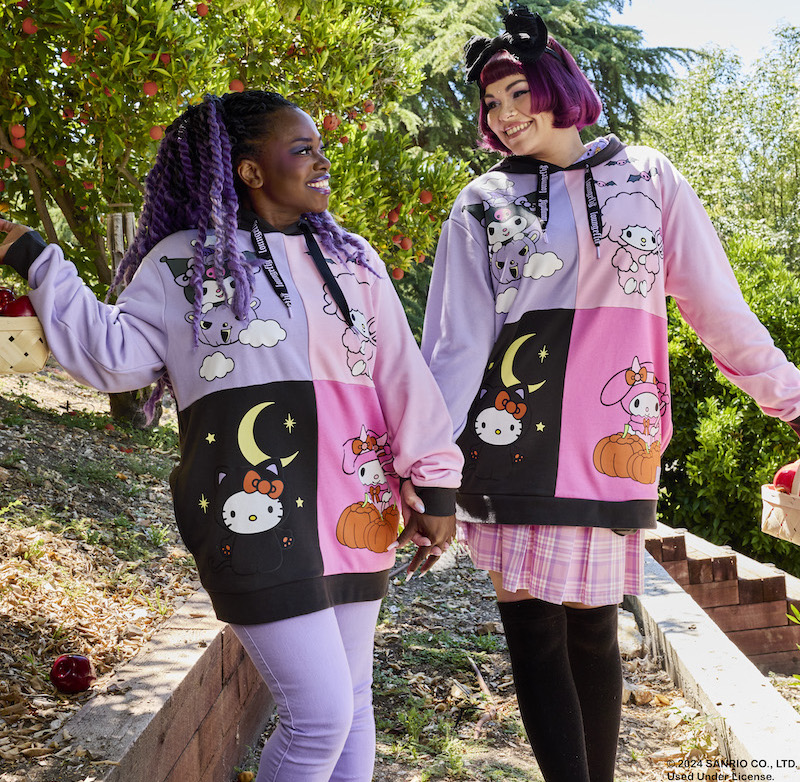 Two women standing outside in an apple orchard holding hands, wearing matching Loungefly Sanrio Hello Kitty and Friends Costume Unisex Hoodies, which feature Hello Kitty, My Melody, Kuromi, and My Sweet Piano dressed in different Halloween costumes in a color-block style.