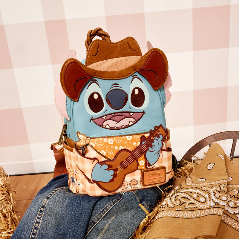 Loungefly Western Stitch mini backpack featuring Stitch dressed in western attire, including an appliqué cowboy hat. The bag sits on a pile of denim and a bandana with a light pink gingham background. 