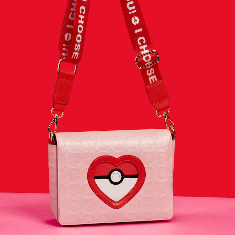 Pink Loungefly Pokémon crossbody bag featuring a heart-shaped Pokéball on the front and a red crossbody strap that says “I Choose You.” The bag sits against a pink and red background. 