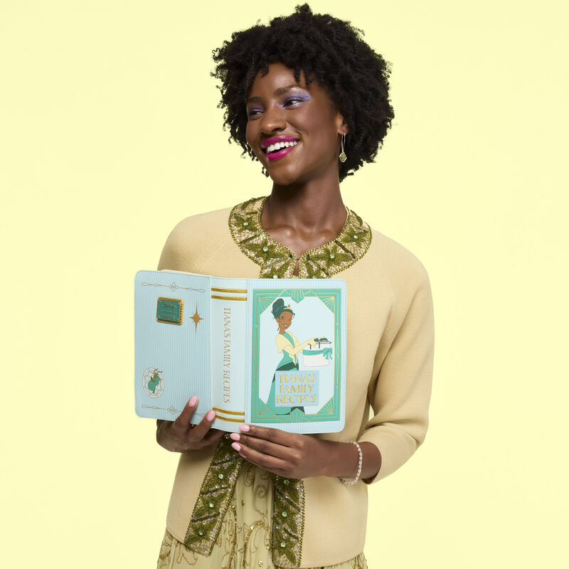 Woman wearing a yellow-toned sweater with green embroidery, standing against a yellow background. She holds the Loungefly Disney Princess Tiana Recipe Book Crossbody Bag in her hands, open like a book. 