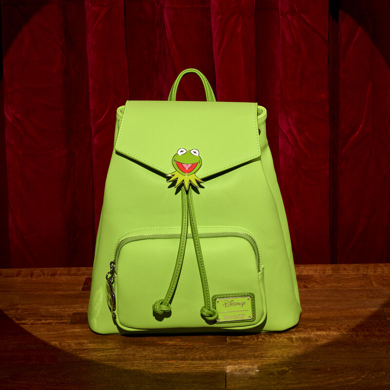 Green Loungefly Kermit mini backpack with drawstrings and a small front pocket. There’s a Kermit the Frog enamel charm on the front flap. It sits on a wooden floor with a red theatre curtain behind it. 