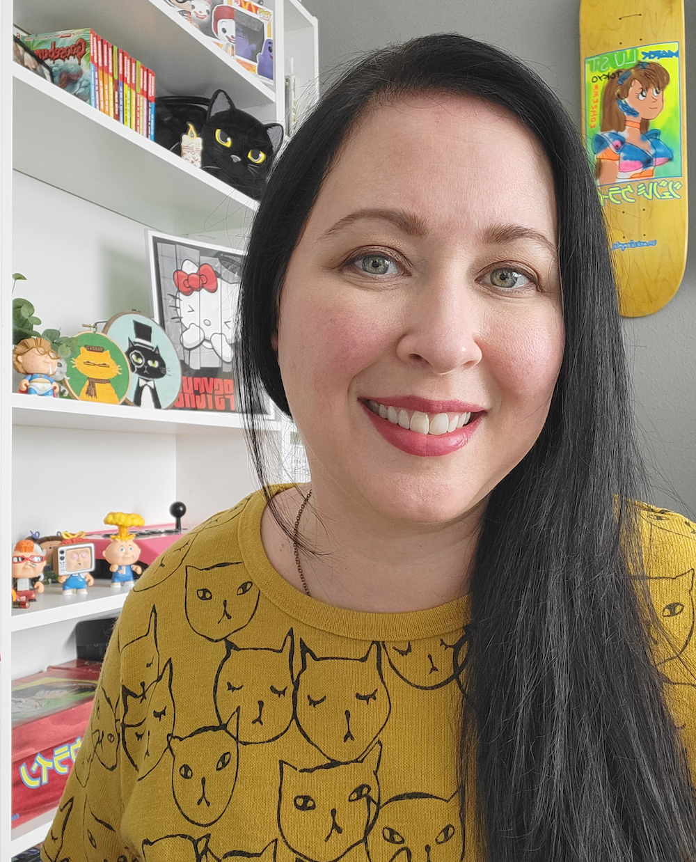 Brunette woman smiling at the camera wearing a yellow sweater with the outlines of cats on it. She sits in front of a bookshelf in an office. 