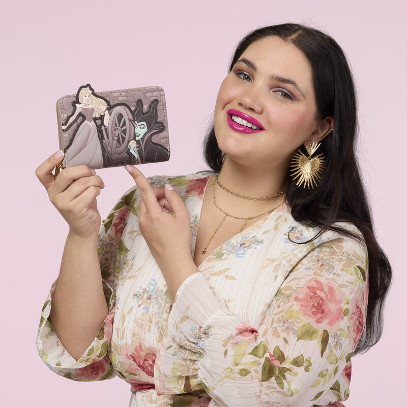 Woman wearing a floral dress, holding the Loungefly Disney Sleeping Beauty Spinning Wheel Zip Around Wallet in one hand.  She stands against a pink background. 