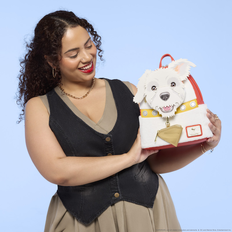 Woman wearing a khaki colored dress with black vest holding the Loungefly Supergirl Krypto mini backpack. 