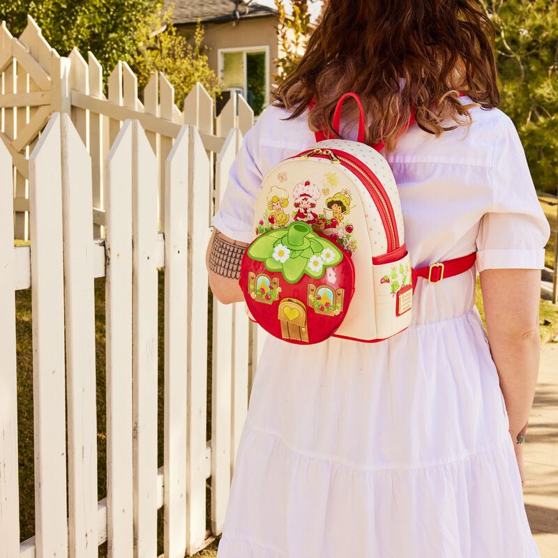 Woman wearing a white dress by a picket fence wearing a white and red backpack featuring Strawberry Shortcake's house and friends