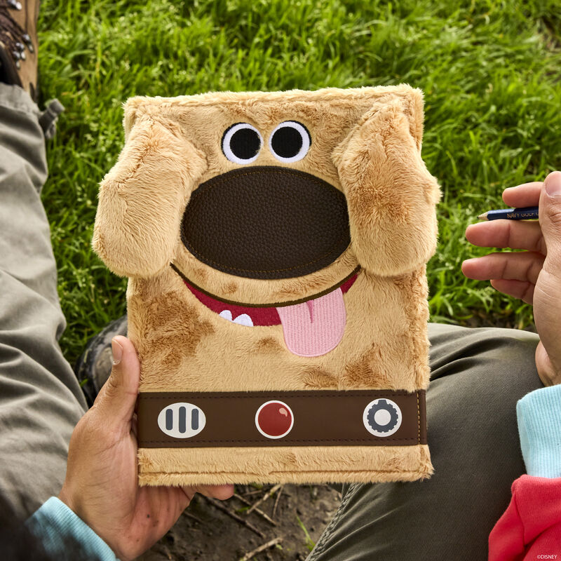 Person sitting outside on the grass and holding the Dug plush journal, featuring 3D ears and embroidered facial details. 