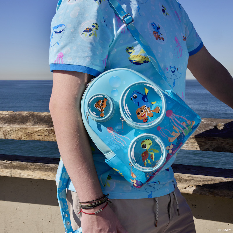 Man at the beach wearing the blue Finding Nemo 20th Anniversary T-Shirt and the Finding Nemo 20th Anniversary Bubble Pocket Mini Backpack, leaning against a wall