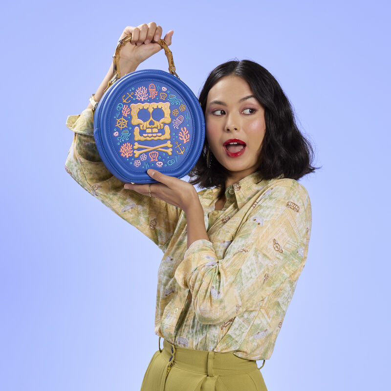Woman holding up the Loungefly SpongeBob SquarePants Pirate Crossbody Bag next to her face. She wears a yellow blouse with an olive green skirt. She stands against a blue background. 