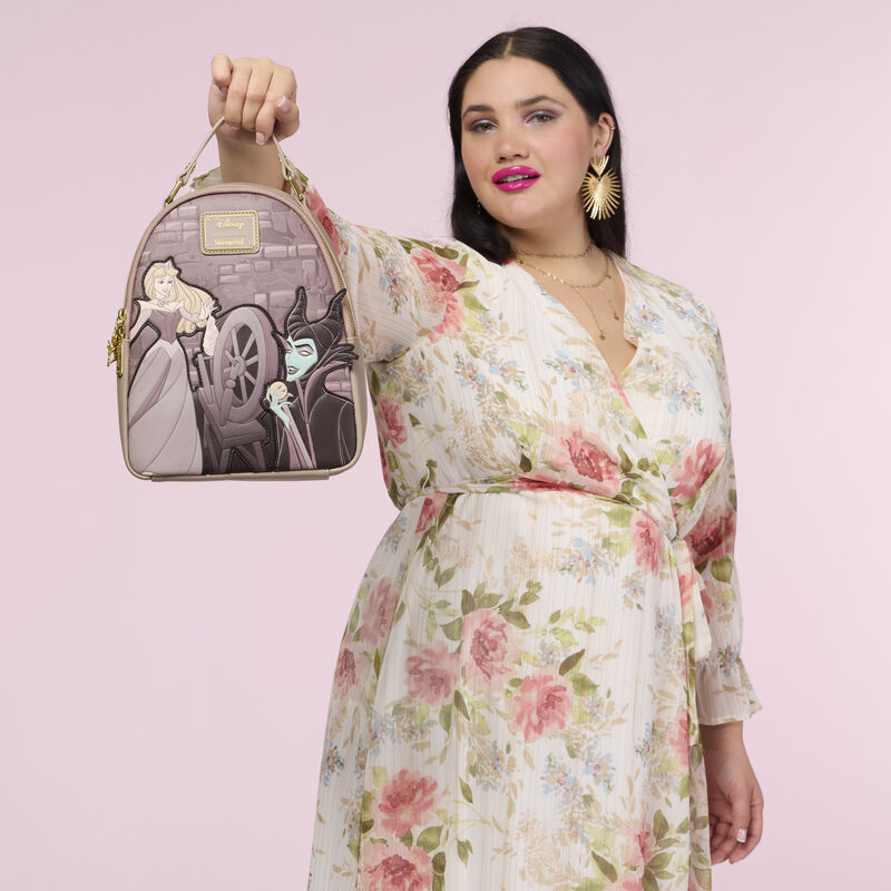 Woman wearing a floral dress standing against a pink background. She holds up the Loungefly Disney Sleeping Beauty Spinning Wheel Mini Backpack by the handle. 