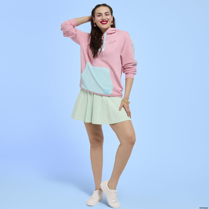 Woman posing in front of camera wearing the Loungefly Barbie hoodie with a green skirt and white shoes. She stands against a blue background. 