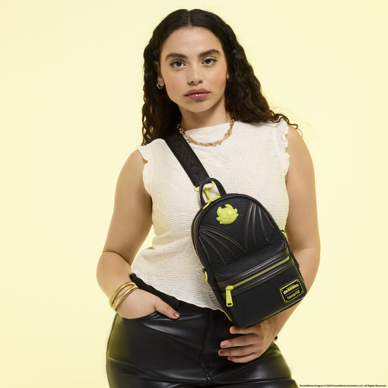 Woman wearing a white sleeveless shirt and black leather pants, wearing the Loungefly How to Train Your Dragon Night Fury Sling. She stands in front of a light yellow background. 
