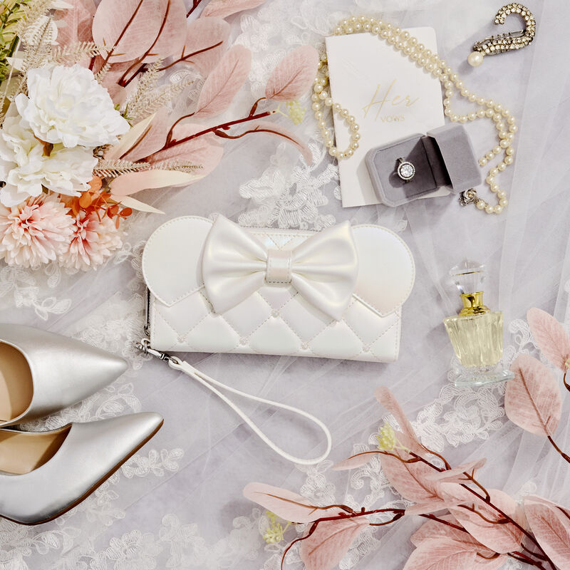 Flat lay of the Loungefly Disney Minnie Mouse Iridescent Wedding Zip Around Wallet laying against a veil surrounded by other wedding-themed items like silver heels, flowers, a pearl necklace, and perfume.
