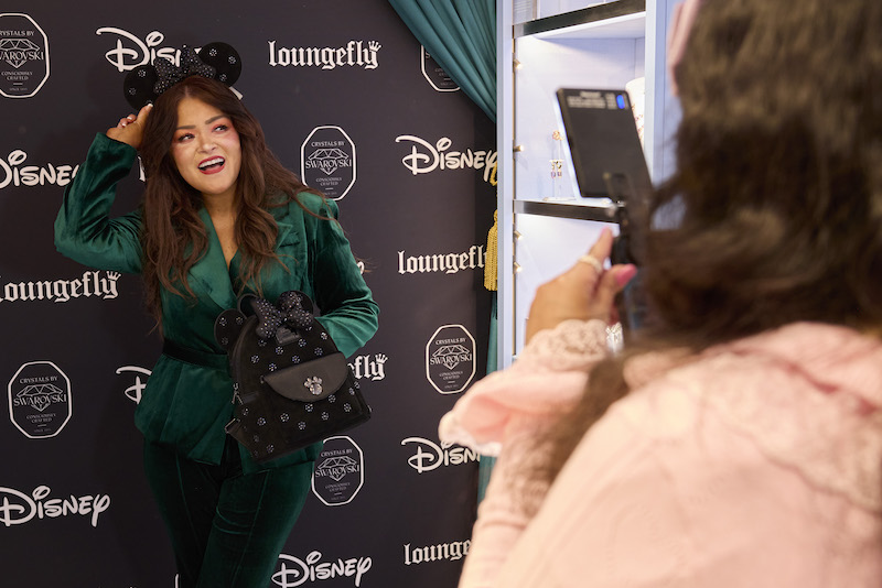 Woman wearing a green ensemble posing in front of the Loungefly x Disney x Swarovski backdrop, posing with the ear headband and mini backpack. 