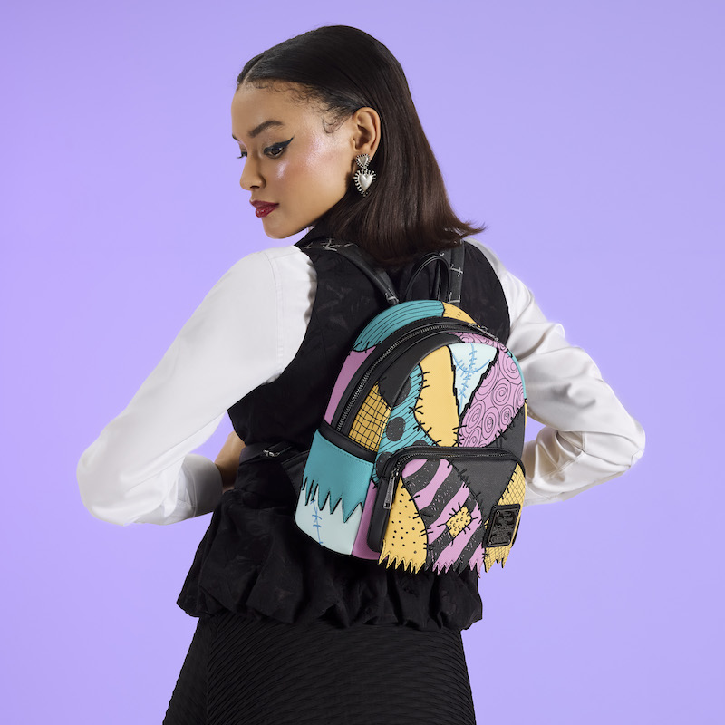 Woman facing away from camera wearing a black vest over a white long-sleeve shirt and a black skirt. She wears the Loungefly Nightmare Before Christmas Sally mini backpack. She stands against a purple background. 