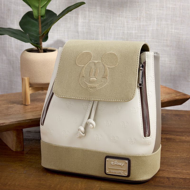 The Mickey and Friends Canvas bag sits on a wooden table beside a green plant against a gray backdrop. 