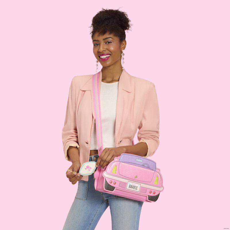 Woman wearing blue jeans, a white top, and a pink blazer. She wears the Loungefly Barbie Corvette Crossbody Bag over a shoulder and showcases the key-shaped coin bag. She stands against a pink background. 