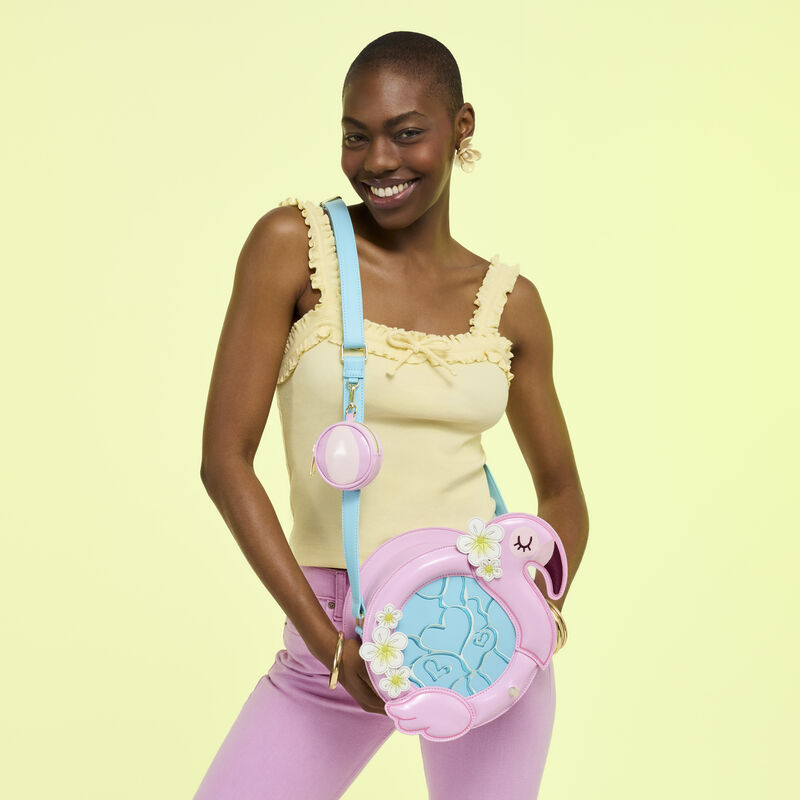 Woman posing in front of camera with the Loungefly Flamingo Pool Float crossbody bag. She wears a yellow tank top and pink jeans and stands in front of a yellow background. 