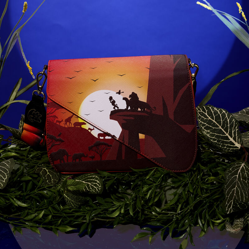 Loungefly Lion King crossbody bag featuring the scene where Rafiki holds Simba up at Pride Rock to show the other animals with the sun in the background. The bag sits amongst vegetation against a dark blue background.