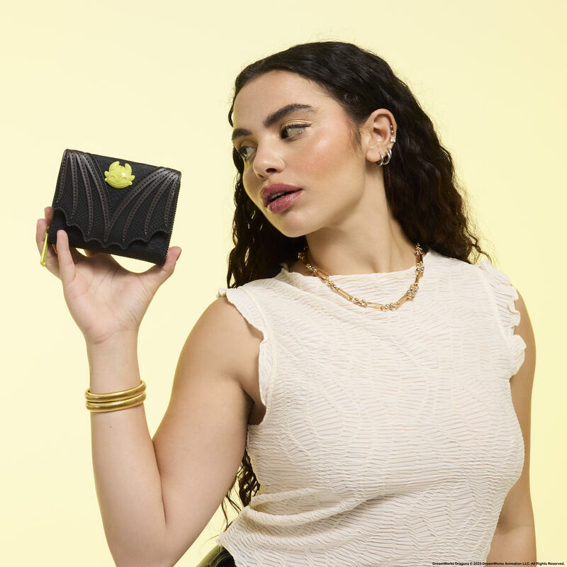 Woman holding up the Loungefly How to Train Your Dragon Night Fury wallet and looking at it. She wears a white sleeveless top, gold bracelets, and a gold necklace. She stands against a light yellow background. 