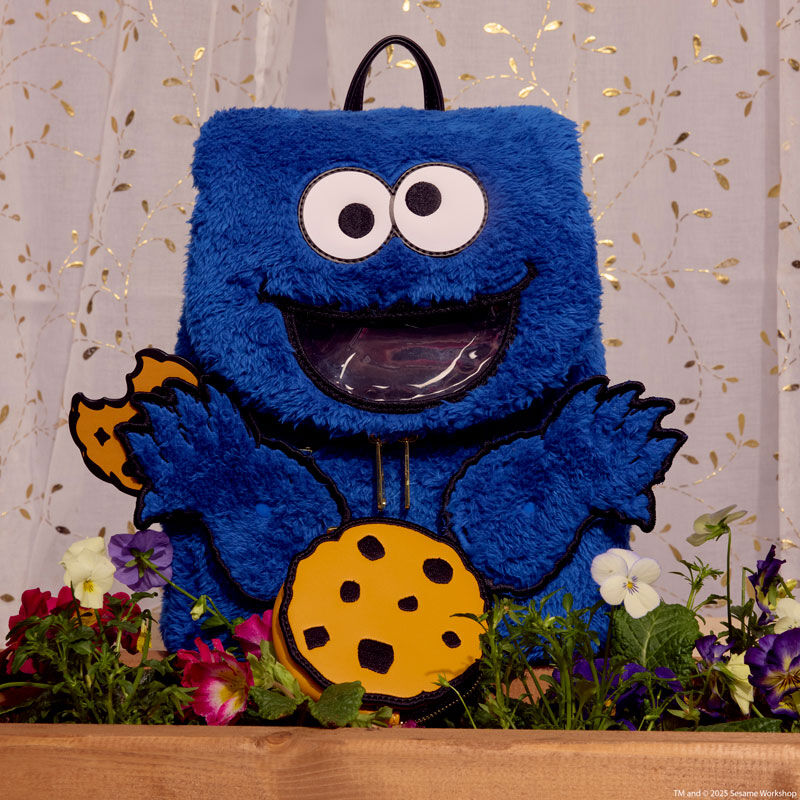 Blue plush Loungefly Cookie Monster mini backpack sitting in a flowerbed in a windowsill with translucent curtains behind it. Cookie Monster holds a half-eaten cookie in one hand, and the cookie coin bag sits in front of the mini backpack. 