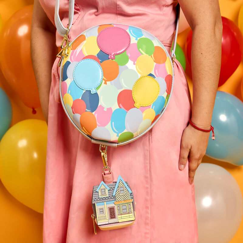 Close-up shot of woman wearing a pink dress and wearing the Loungefly Up Balloon House Figural Crossbody Bag, which looks like the bunch of balloons Carl uses to float his house on the main compartment. The coin bag dangles below, which is in the shape of Carl’s house. 