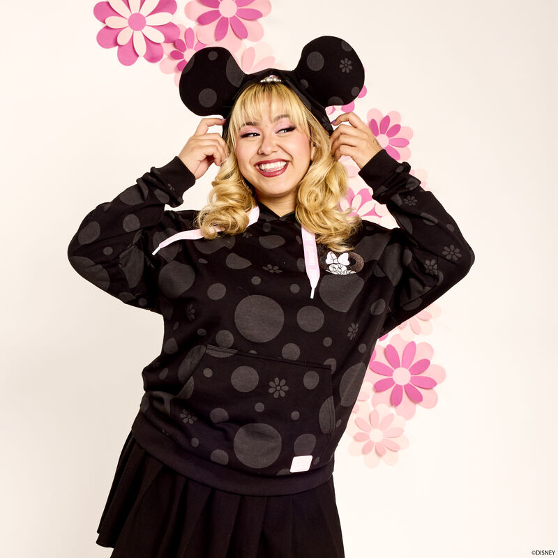 Blonde woman smiling and wearing the black Loungefly Minnie Mouse Floral Rock the Dots hoodie with the hood up to show off the Minnie Mouse ears. She stands in front of a white background with a pink floral design on it. 