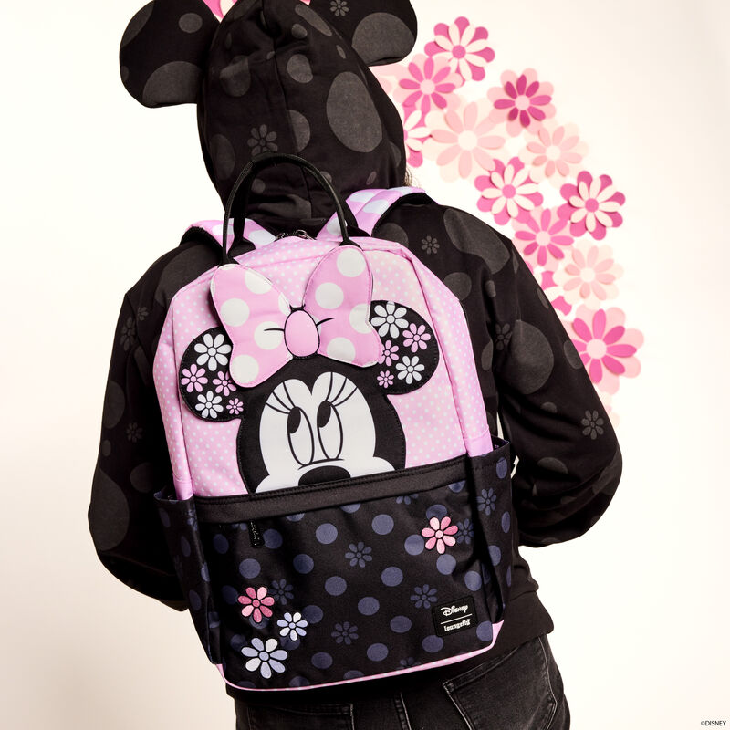 Woman facing away from camera wearing the black Loungefly Minnie Mouse Floral Rock the Dots Hoodie with the hood up and wearing the nylon Loungefly Minnie Mouse Floral Rock the Dots Full-Size Backpack. She stands in front of a white background with pink floral designs on it. 