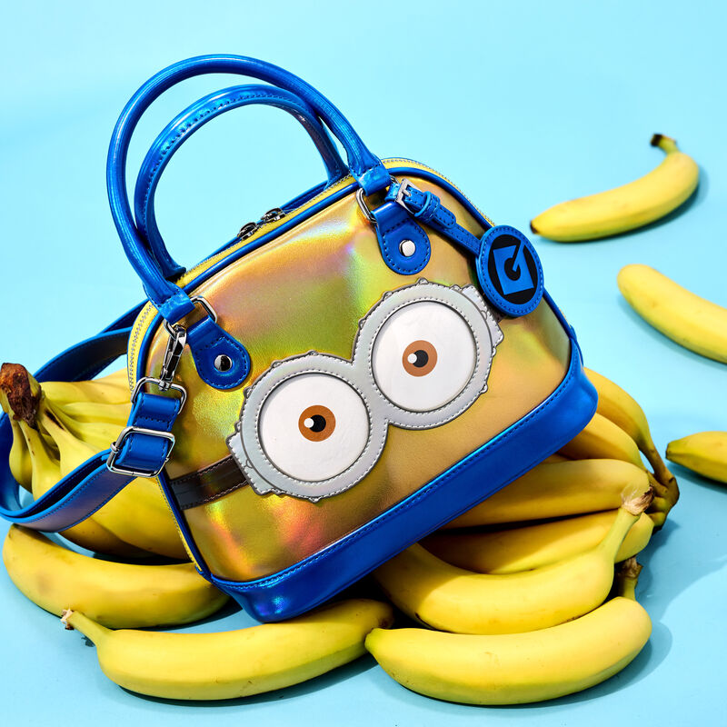 Domed Loungefly crossbody that's iridescent yellow and blue with the design of Minion from Despicable Me on it sitting on a bunch of bananas against a light blue background