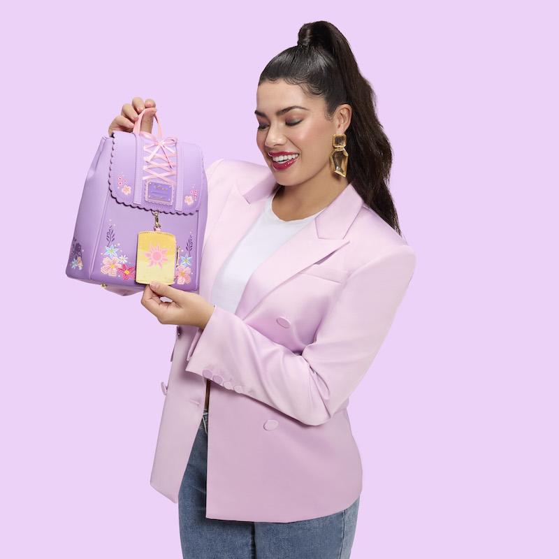 Woman wearing blue jeans, a white top, and a purple blazer holding the exclusive Loungefly Disney Tangled Rapunzel Mini Backpack. She stands against a purple background. 