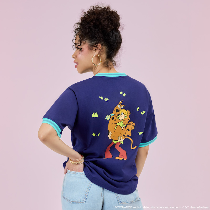 Woman facing away from camera with a hand in the back pocket of her light-wash denim jeans and wearing the Loungefly Scooby-Doo Ringer Tee, featuring Scooby and Shaggy on the back, surrounded by pairs of spooky eyes. She stands in front of a pink background. 
