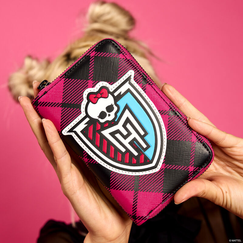 Woman holding the pink plaid Loungefly Monster High zip around wallet to the camera. The front features the Monster High crest. The woman stands in front of a pink background. 