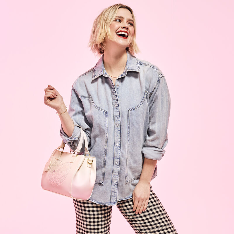 Woman looking off to the side and smiling. She wears an oversized denim button-up with black-and-white checkered pants. She carries the Loungefly Stitch and Angel Cherry Blossom Heart Crossbody Bag over one arm. She stands against a pink background.