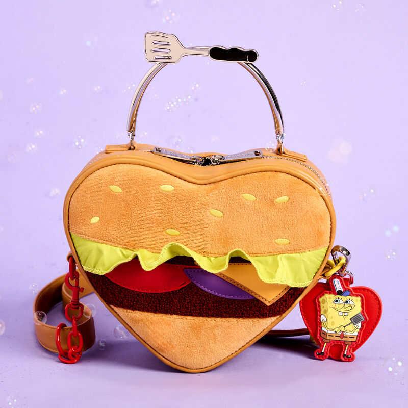 Heart-shaped plush Loungefly crossbody in the shape of a Krabby Patty from SpongeBob SquarePants. The bag sits against a purple background with bubbles floating around it. 