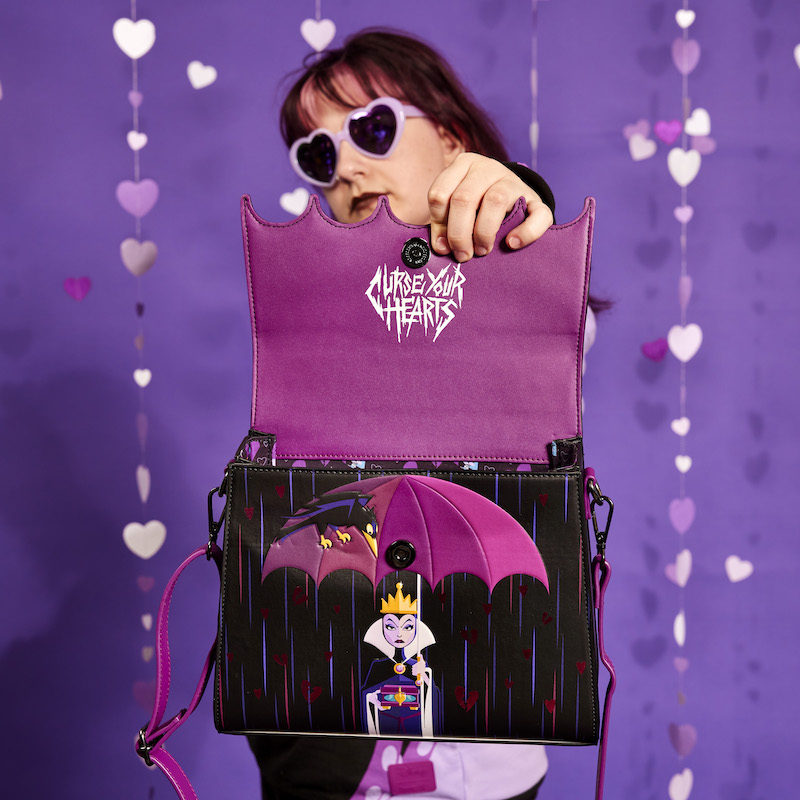 Image of woman holding the Loungefly Disney Villains Curse Your Hearts Crossbody Bag out to camera, holding the top flap up to reveal "Curse Your Hearts" written underneath and the Evil Queen's Raven nestled in her umbrella 