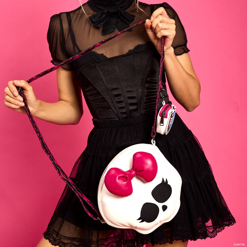 Woman wearing a vintage-style black dress standing against a pink background and holding the figural Skullette crossbody bag, featuring Skullette on the front with a 3D bow and a Monster High crest coin pouch attached. 