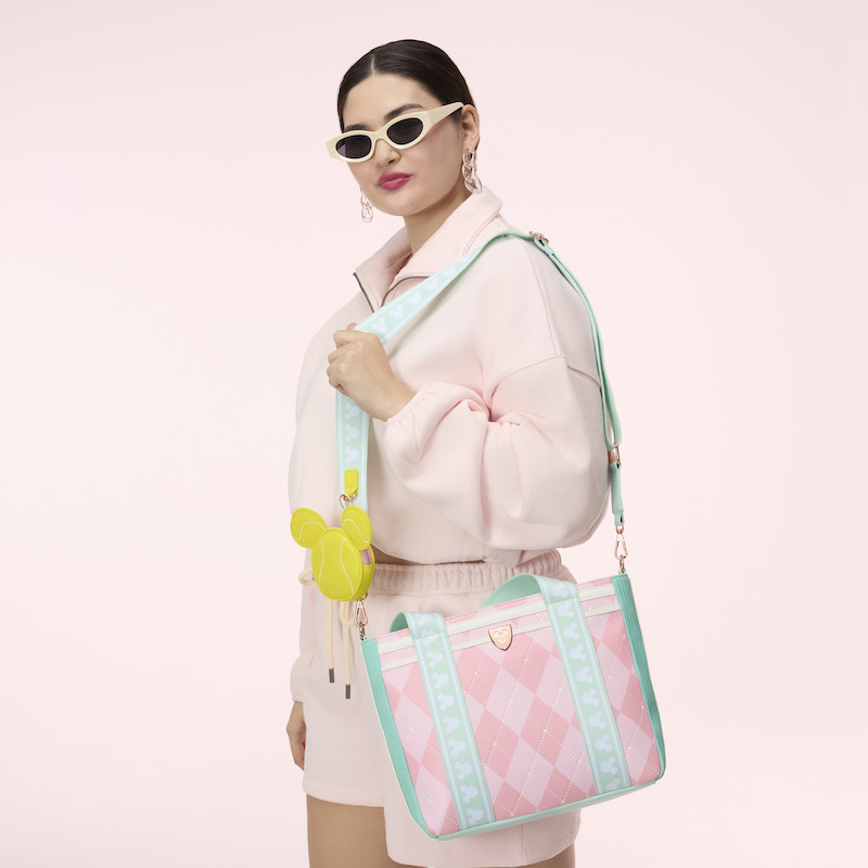 Woman wearing pink shorts and sweater and sunglasses, wearing the Loungefly Disney Mickey Mouse Tennis Tote over a shoulder. She stands against a pink background. 