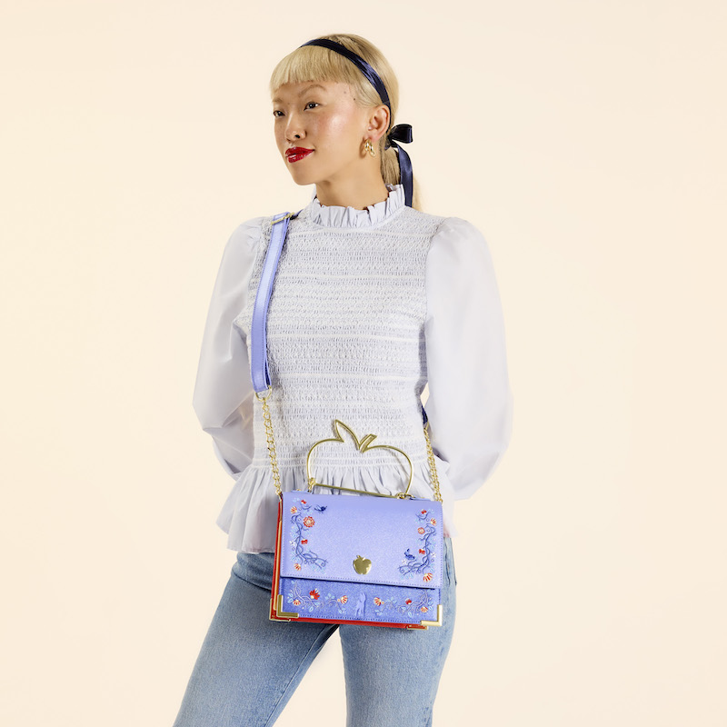 Woman wearing a light purple blouse and blue jeans, wearing the Loungefly Disney Princess Snow White crossbody bag. She stands in front of a light pink/beige background. 