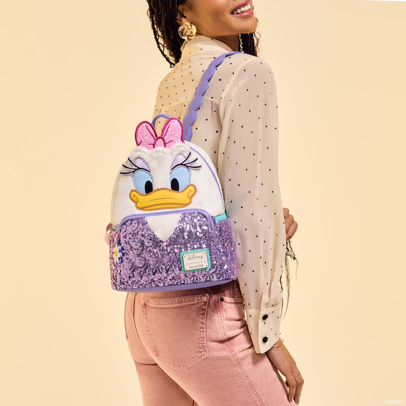 Woman facing side-profile to camera, wearing a sheer neutral black-polka-dot blouse and brick-colored jeans. She wears the plush and sequin Loungefly Daisy Duck mini backpack. She stands against a beige background.