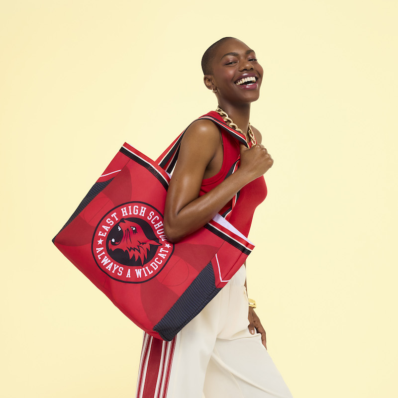 Woman smiling at camera wearing a red top and white pants with red stripes, wearing the Loungefly High School Musical Wildcats Canvas Tote Bag over a shoulder. She stands in front of a yellow background. 