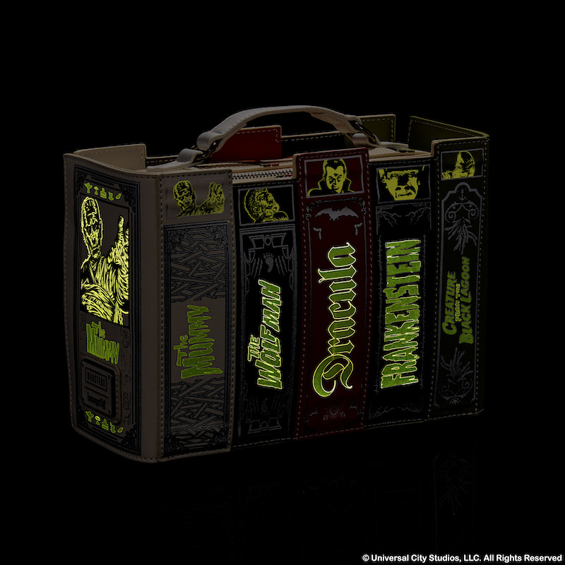 Loungefly Universal Monsters Books Glow Crossbody bag sitting against a dark background in the dark to show off the glow in the dark details of the titles