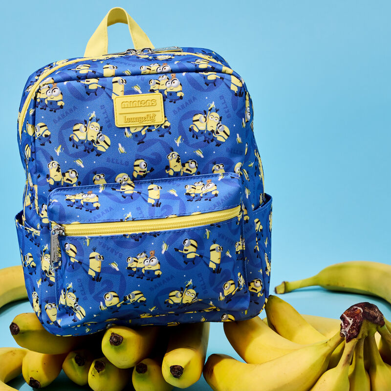 Square nylon mini backpack featuring Minions characters and bananas all over a blue background. The bag sits on a pile of bananas against a blue background.