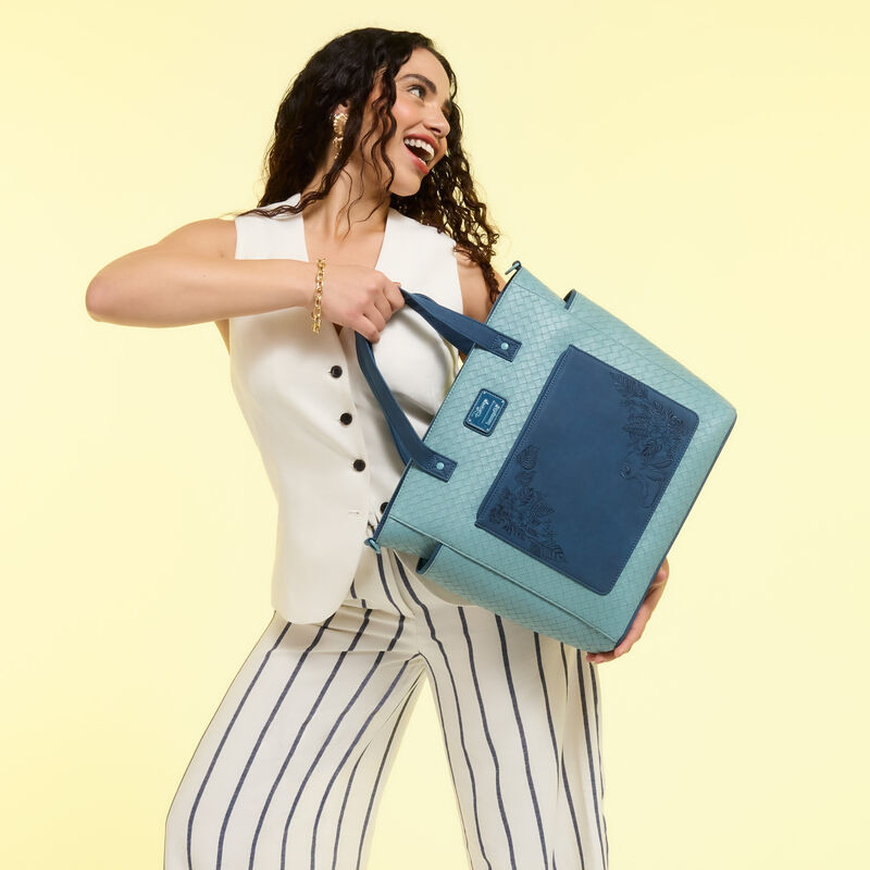 Woman wearing a button-down white blouse with white-and-black striped trousers. She holds the blue Loungefly Stitch Embroidered Floral Convertible Tote on an angle as she looks to the side and smiles. 