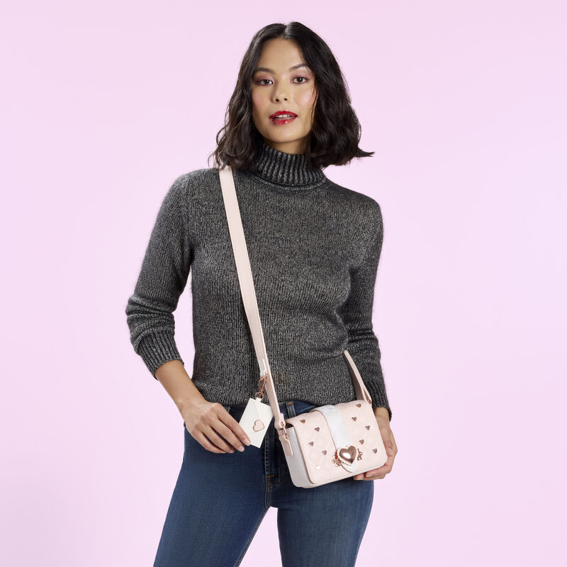 Woman wearing a grey turtleneck sweater with blue jeans, wearing the Loungefly Valentine&rsquo;s Day Heart Envelope Crossbody. She stands against a light pink-purple background. 