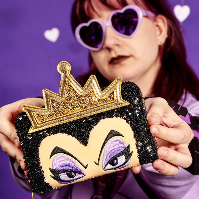 Woman wearing Purple Heart-shaped sunglasses holding the sequin Evil Queen zip around wallet, featuring the Evil Queen on the front toward the camera. She stands against a purple background. 